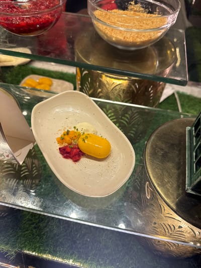 A white, oblong plate contains a dessert: a bright yellow, capsule-shaped component, accompanied by a dollop of white cream. Small heaps of diced strawberries and mango segments are arranged near the capsule. Sprinkle of chopped pistachios and biscuit crumbs garnish the plate. Behind the plate, there's a display of ingredients, including a jar of biscuit crumbs and another filled with what appears to be diced strawberries. The scene is reflected in a glass surface.