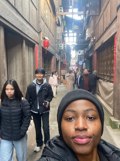 A selfie with three young people in a wooden structure setting. The background features traditional Chinese architecture with red lanterns, aged wood paneling, and a bustling crowd of people. The foreground focuses on the individuals: a person with a beanie, followed by two other young individuals with dark hair and jackets. The environment suggests a historical or cultural site.