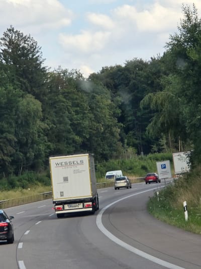 The image shows a view from a vehicle on a highway, likely in Germany, given the signage and truck manufacturer. A white semi-trailer truck with "WESSELS LOGISTIK" visible on its side is in the foreground, traveling on a two-lane highway. The truck's license plate reads "B-DL 197" and "SCHMITZ". To the left of the truck, a dark-colored car is partially visible, also on the highway. Further ahead, a red car and a silver car are visible in the right lane. A white van is also present in the distance. A white sign with a blue "P" symbol and German text, likely indicating a parking area, is visible on the right side of the road. The highway is bordered by dense green trees and vegetation on both sides. The sky is partly cloudy. The location is identified as Ellenhausen, Germany, due to the presence of a sign for "Parkplatz Welchenfahrn", which is an area within Ellenhausen.