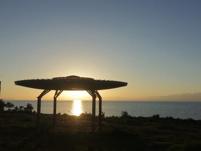 A silhouetted UFO-shaped pavilion stands in the foreground, its wide, flat roof supported by four dark pillars. The pavilion casts a shadow over a grassy, uneven landscape in the foreground. Beyond the pavilion, a vast body of water, likely Lake Issyk-Kul, stretches towards a distant, hazy shoreline with mountains on the horizon. The setting sun is visible low in the sky, casting a bright, shimmering reflection on the water's surface. The sky transitions from a pale blue at the top to a warm yellow and orange hue near the horizon. The image is a photograph taken from a slight elevation, looking out towards the lake during sunset.