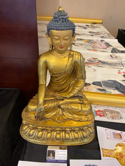 The image shows a golden statue of a reclining Buddha, possibly made of bronze, resting on a black surface. The statue has blue, curly hair and closed eyes, with its head tilted to the side. A golden robe is draped over its body, revealing intricate folds and details. Behind the statue, a large, colorful tapestry or scroll depicts figures in traditional attire, possibly from a historical or religious context. There are also several printed images of artifacts, some with Chinese text, displayed on the wall behind. One of these images is a catalog entry with "2026香港皇家珍藏藝術品拍賣會" (2026 Hong Kong Royal Collectible Art Auction) visible. The overall setting appears to be an exhibition or auction preview, given the displays and the presence of catalog information. The lighting suggests an indoor environment. There are no overt indicators of weather or time of day, but the atmosphere seems serene, fitting for a religious artifact. The location context provided is Futian District, China, which aligns with the presence of Chinese text and the nature of the displayed items.