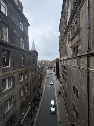 The image shows a view from above, looking down a narrow, wet street lined with old stone buildings in the city of Edinburgh, United Kingdom. The architecture is predominantly old, with many windows and some decorative stonework visible on the facades. The street is paved and has double yellow lines marking the center. Several cars are driving or parked on the street, and a few pedestrians are walking along the sidewalks. The sky is overcast and gray, contributing to a somber atmosphere. In the background, more buildings with similar architectural styles are visible, along with a distinctive church spire. The scene appears to be a typical urban street in an older part of the city.