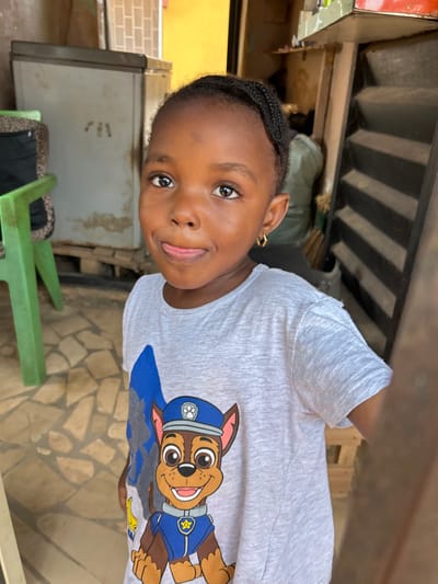 A young child with dark skin, dark braided hair, and gold-colored hoop earrings is centered, looking directly at the camera with a playful expression, extending their tongue slightly. They are wearing a light grey short-sleeved t-shirt with a blue and brown graphic of the Paw Patrol character "Chase" in a police uniform. To the child's left, a green plastic chair is partially visible in the foreground, with an older, light-colored chest freezer behind it. The floor is covered with light-colored, irregularly shaped tiles. To the child's right, a dark, shadowed doorway or opening with vertical slats is present, revealing a glimpse of a yellow wall and a dark object or person's limb in the background. The scene takes place in Ashipa, Nigeria.