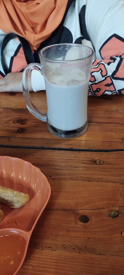 The image shows a glass mug filled with a light-colored beverage, likely milk or a milk-based drink, sitting on a wooden table. The mug has a handle and is made of clear glass. In the background, an arm is visible, wearing a white shirt with orange accents and a black strap on the wrist. The table is brown and wooden, with a grain pattern. A partially visible orange plate with a brown, cylindrical object on it is in the foreground. The scene appears to be indoors, possibly at a cafe or restaurant in Mojokerto, Indonesia, at the coordinates provided. The scene is captured from a high angle, with a neutral lighting condition. There is no apparent interaction occurring between the subjects in the scene.