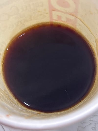 The image depicts a close-up, overhead view of a dark, deep brown liquid, consistent with coffee, contained within a white, disposable paper cup. The liquid fills the majority of the frame, exhibiting a reflective surface and a distinct meniscus where it meets the cup's interior wall. Small, light-colored air bubbles are present along the meniscus, particularly on the left side. Faint branding is visible on the inner wall of the cup, above the liquid, including partial, reddish-brown text that appears to spell "COFFEE CO" and potentially "LAVAZZA". A few minuscule white particulate specks are discernible on the liquid surface. The scene is located in the city of Lehvaz, Armenia. No individuals or discernible actions are present.