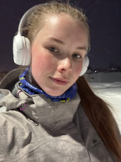 A young woman wearing white headphones and a blue patterned neck gaiter poses for a selfie. She is wearing a gray jacket. The background is dark, possibly nighttime, with a suggestion of snow or a light-colored structure. There is nothing present in the image to confirm the location as the city of Berezniki, Russia.