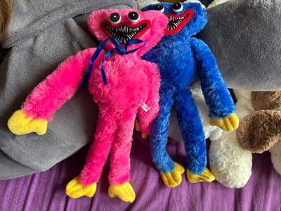 Two plush toys, one pink and one blue, are positioned on a purple, textured surface. The pink toy is on the left and the blue toy is on the right. Both toys have large, circular eyes, sharp-toothed smiles with red lips, and yellow feet. A blue ribbon is tied around the neck of the pink toy. The toys appear to be lying on top of gray, textured fabric. The background is blurred but suggests a domestic setting. The location in the city of Парэчча, Belarus, is not discernible from the image.
