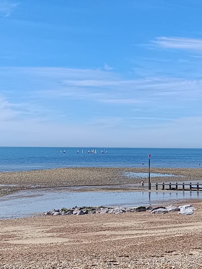 The image depicts a beach scene with the sea in the background. The sky is a clear, bright blue with a few wispy white clouds. The sea is a deep blue and appears calm. In the distance, several small sailboats are visible on the water.

The foreground of the image is a beach covered in small pebbles and sand. There is a tidal pool or shallow water area in the mid-ground, with rocks arranged along its edge. A wooden structure, likely a groyne, extends into the water from the right side of the frame. A single person is walking along the wet sand near the groyne, and a red-topped post stands vertically on the beach. The overall impression is a peaceful seaside environment, likely in Arun, United Kingdom, given the typical coastal scenery.