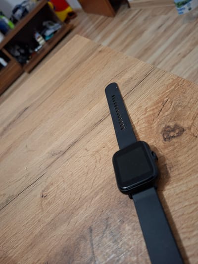 A black smartwatch with a black band lies on a light brown wooden table. The watch face is dark and rectangular, and the band has several small holes for adjustment. The wood grain is visible on the table surface, showing variations in color and texture. In the blurred background, a dark brown wooden shelf unit is visible, containing various objects, some of which appear to be containers and miscellaneous items. The overall scene suggests a domestic or indoor setting, possibly a home in Ilava, Slovakia, given the user's prompt.