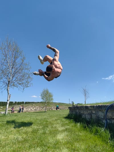 A shirtless, muscular man with tattoos is in mid-air performing a backflip. He is wearing dark shorts and white sneakers. The background is a grassy field with a few trees and a blue sky. A stone wall is visible in the lower right corner. In the distance, there are people near some trees. The location is Neofit Rilski, Bulgaria.