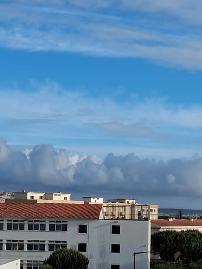 The image presents a vertical view of a scene in Figueira da Foz, Portugal. On the right, buildings are visible, with light-colored facades, some featuring red-tiled roofs and windows. The urban environment includes greenery in the form of trees or bushes. The left side of the image is dominated by the sky, featuring a mix of blue sky and clouds. The clouds vary in density, ranging from thin, wispy formations to denser, darker cumuliform clouds.