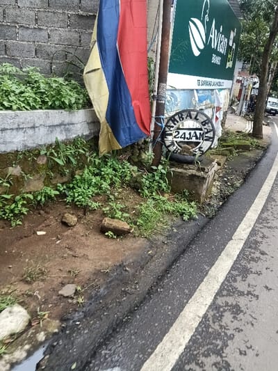 A street scene showcases a sidewalk bordering a gray brick wall and a road with a white line marking the edge. Vegetation covers the wall's base. A flag with yellow, blue, and red stripes hangs near a pole, next to a sign for "Avian Brands." A round sign reading "24 JAM" sits atop a small concrete structure. The ground near the sidewalk appears muddy and littered with debris. In the background, cars are visible on the road.