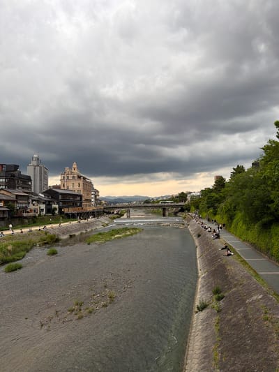 This image depicts a wide view of the Kamo River (Kamo-gawa) in Kyoto, Japan, under a heavily overcast sky. The upper two-thirds of the frame are dominated by a dense, dark grey cloud cover, with a narrow band of lighter, yellowish-grey sky visible low on the horizon in the background.

The Kamo River flows from the background towards the foreground, occupying the lower third of the image. The water is a muted greenish-grey, with visible ripples and a shallow, rocky riverbed, particularly in the middle distance where small clusters of green vegetation grow directly from the riverbed.

On the left bank (west bank), a paved pedestrian path runs parallel to the river. Between the path and the river's edge is a sloped, grassy embankment where approximately 20-30 individuals are seated or walking. Beyond the path, a dense array of buildings lines the river. Closer to the foreground are several traditional-style Japanese buildings with dark roofs and visible wooden elements. In the mid-ground, a prominent, ornate, light brown multi-story building with a distinctive domed roof and classical architectural features stands out. Further back, taller, more modern grey buildings are visible. A multi-arched concrete bridge spans the river in the far distance on this side.

On the right bank (east bank), a steep concrete retaining wall descends to the river. Above this wall, a paved pedestrian pathway runs parallel to the river. Approximately 25-35 individuals are scattered along this path and on the sloped, grassy verge next to the concrete wall, mostly seated. Behind the pathway, a continuous and dense wall of dark green trees and shrubs forms a natural barrier, obscuring any structures beyond it. The same multi-arched concrete bridge is visible in the mid-ground, connecting to this bank.

The people visible on both banks are casually dressed and appear to be engaged in leisure activities such as relaxing by the river. The overall scene suggests a late afternoon or early evening setting due to the low light and cloud conditions.