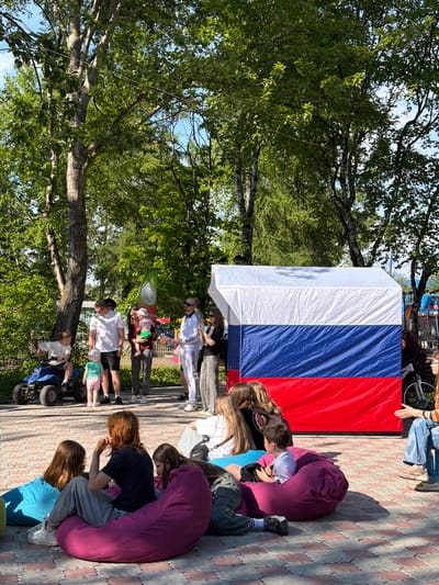 The image captures a lively outdoor gathering, likely a community event or celebration, in Yelizovo, Russia. A large Russian flag, a prominent object, serves as a backdrop, its white, blue, and red stripes stretching across a significant portion of the frame. Several people, including adults and children, are scattered around the flag. Some are seated on large, plush beanbag chairs in shades of purple and blue, while others stand or kneel, interacting with each other.

In the foreground, a child sits on a blue ATV, appearing ready for a ride. Further back, a group of people are gathered around a table with a green flag featuring a white symbol. The presence of trees with lush green foliage suggests a warm, sunny day, likely during the spring or summer. The ground is paved with light-colored bricks arranged in a decorative pattern.

The overall atmosphere seems relaxed and joyful, with people of various ages enjoying the outdoors and each other's company. The clothing worn by the individuals, including t-shirts, shorts, and casual pants, further indicates pleasant weather. The scene appears to be set in a park or an open recreational area. There is no discernible text visible in the image.