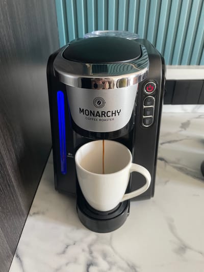 A black and silver Monarchy Coffee Roaster single-serve coffee machine dispenses a dark brown liquid into a white ceramic mug, positioned on a black drip tray. The machine's front panel is silver, featuring the "MONARCHY COFFEE ROASTER" logo and a coffee bean icon. A glowing red power button is visible on the right side, above three smaller buttons, each depicting a coffee cup icon. On the left side, a vertical blue illuminated strip indicates the water level, with a "Min" mark visible near the bottom. The coffee maker rests on a white countertop with a grey marble pattern. To the immediate left of the machine is a dark, possibly wooden, vertical surface. The background features a teal-colored wall with distinct vertical ridges or slats. The scene is situated in the city of Kayuambon, Indonesia.
