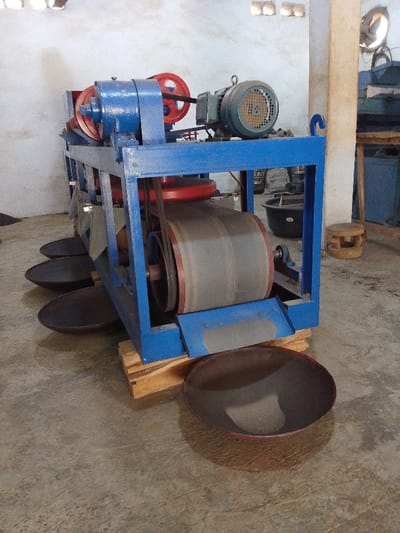 The image depicts an indoor industrial setting within Olomoda, Nigeria, featuring a large, blue-painted processing machine. The machine, constructed with a square-tube metal frame, is equipped with multiple red pulleys, black drive belts, and large, dark grey cylindrical rollers. An electric motor, with a green casing and a protective grille, is mounted atop the frame, connected via a belt to the primary drive mechanism. The machine's base rests on several stacked wooden planks or pallets. Positioned on the concrete floor directly beneath the machine's lower sections are at least five dark, shallow, circular metal pans or trays, likely intended to collect processed material. The background reveals light-colored walls, with three rectangular openings high on the left wall and a vertical support column on the right. Other background elements include a small, light-colored wooden stool, a dark tub or container, and indistinct objects further back. No individuals are present in the scene, and the machine appears to be stationary. The floor is a dusty, light grey concrete.