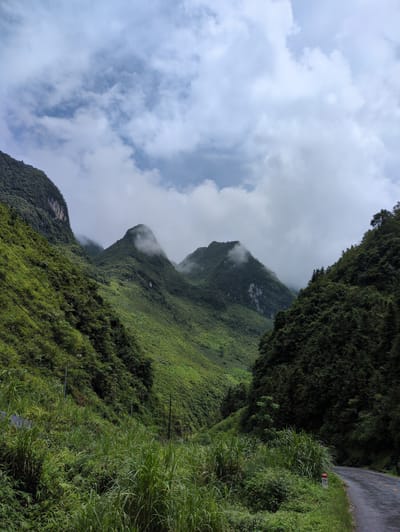 A scenic view of a winding road passing through a lush green valley with dramatic, mist-shrouded mountains under a cloudy sky. The road, visible on the right side of the frame, is paved and appears wet, suggesting recent rain. Tall, dense vegetation, including grasses and trees, flanks the road and covers the steep mountain slopes. Power lines stretch diagonally across the left side of the image, traversing the mountainside. A small, red and white marker post with the inscription "H4 110" stands near the roadside on the right. The mountains are characterized by their steep inclines and varied textures, with some peaks partially obscured by clouds. The overall impression is one of natural beauty and remoteness. The scene is consistent with the mountainous terrain found in Phố Bảng Commune, Vietnam, known for its dramatic landscapes. There are no people or animals visible in the image.