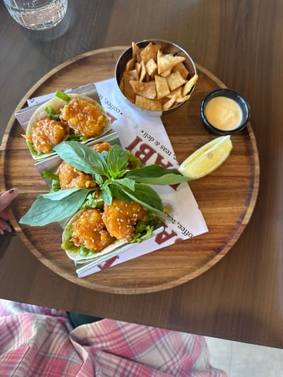 An overhead shot displays a meal set on a circular wooden tray, which rests on a darker wooden table. The centerpiece of the meal is three small tacos, each featuring crispy, fried protein (possibly shrimp or chicken) coated in a glistening orange sauce, sprinkled with sesame seeds, and nestled on a bed of fresh lettuce. A vibrant sprig of green basil adorns the central taco.

Beside the tacos, a small metal bowl holds a generous portion of light brown tortilla chips. A black ramekin contains a creamy, light yellow dipping sauce, and a fresh lemon wedge with a visible seed is placed nearby. The tacos are supported by metal holders lined with white paper.

A person, visible on the right, wears a pink and white plaid shirt, with their hand featuring a manicured nail lightly touching the tray area. Part of a ridged glass can be seen on the far left. Visible text on the paper lining beneath the tacos includes "LABI" and phrases like "Coffee, teas & deli," indicating a casual dining setting, possibly a cafe or deli. The bright, natural lighting suggests it is daytime. The scene captures a moment of dining in Polje, Montenegro.