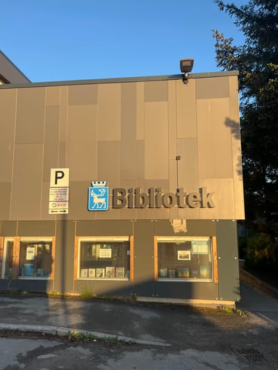 The image shows the exterior of a library building in Tromsø, Norway, on a sunny day. The building has a modern facade with large windows displaying books. A prominent sign above the windows reads "Bibliotek" (library) in large, gray, three-dimensional letters. To the left of the sign is a blue and white emblem featuring a reindeer, the symbol of Tromsø. Below the emblem and to the left of the emblem are parking signs. The building is surrounded by asphalt pavement and a small patch of grass. In the background, a large coniferous tree is visible. The sky is clear and blue. The lighting suggests it is late afternoon, with the sun casting long shadows.