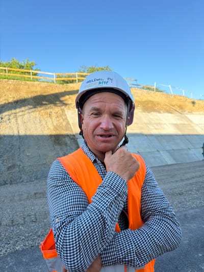 A man in a construction setting is captured in this slightly tilted, medium shot. He wears a white hard hat with blue lettering that reads "WELDTech" and some illegible text, along with an orange safety vest over a black and white checkered button-down shirt. His right hand is near his neck, and he has a slight smile on his face, conveying a relaxed or perhaps amused emotion.

The background suggests an outdoor construction site. A light-colored, rough-textured wall or structure is visible behind him, with shadows indicating sunlight. To the left, the bright blue of a clear sky is prominent. A wooden fence with white posts runs along a dirt or gravel path in the mid-ground, with some greenery and trees visible beyond. The presence of the hard hat and safety vest clearly indicates a work environment, likely on a construction project. The lighting and clear blue sky suggest it is daytime, and the shadows indicate it could be late morning or early afternoon. The location context provided is Jodłownik, Poland.