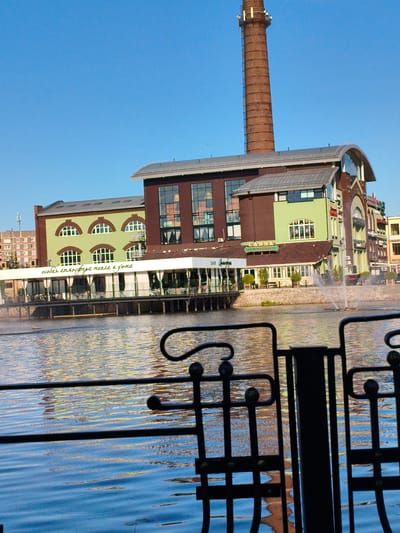 The image shows a multi-story building complex with a prominent brick chimney, situated next to a body of water. The building has a mix of architectural styles, featuring large windows, arched windows, and a curved roof section. One section of the building is painted green, and another is brick red. A white awning extends over a section of the building that appears to be a restaurant or cafe, with seating visible inside. There is a fountain in the water in front of the building. A black metal fence is in the foreground, partially obscuring the view of the water and the building. The sky is clear and blue. The location is identified as the former Klyuch factory in Noginsk, Russia. No people are clearly visible.