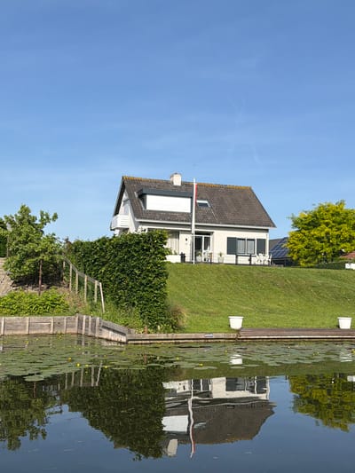 The image shows a house and its reflection in water. The house is white with a dark gray roof and has a flag pole with a flag on top. There's greenery around the house, including a large bush and trees. The water reflects the house, the trees, and the sky. Lily pads with yellow flowers are floating on the surface of the water near the bank. The sky is blue with a few wispy clouds. The location is Rhenoy, Netherlands.