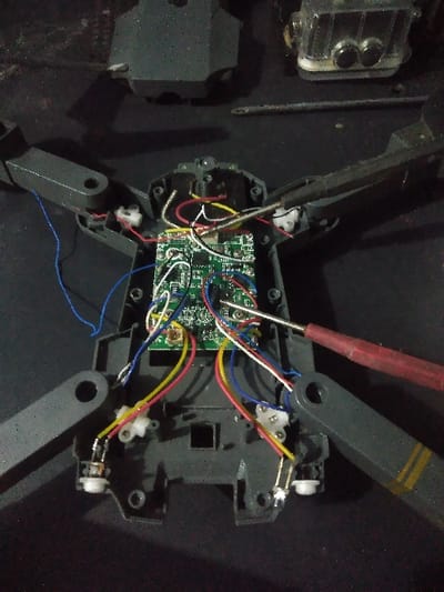 The image displays a partially disassembled grey quadcopter drone, possibly a DJI Mavic Pro series model, laid open on a dark surface, consistent with a workbench. The top casing of the drone has been removed, revealing a central green printed circuit board (PCB) to which numerous multi-colored wires (red, yellow, blue, white, black) are connected. These wires extend to various points within the drone's grey plastic chassis, including small white connector blocks near the points where the arms attach. The drone's four foldable arms are in an extended position, with one arm on the right featuring yellow and black striped tape near its end. A screwdriver with a red handle and silver shaft is positioned on the right, its tip near the internal circuitry, suggesting ongoing repair or assembly work. In the upper background, to the right, a clear or translucent plastic waterproof housing, resembling an action camera case with two dark circular elements, is visible. Another dark grey drone component, likely part of its casing, is in the upper left background, along with a thin silver rod-like tool. No individuals are present. The location in the city of Portland North, Australia, cannot be determined from the visual information provided.