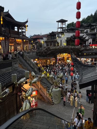 The image depicts a crowded street scene in Zhangjiajie, China. Traditional Chinese buildings line both sides of the street, illuminated by lanterns and lights. A large crowd of people is walking through the area. Red lanterns are strung overhead, while other lanterns hang from buildings. A large, gnarled tree stands prominently near the center of the scene.