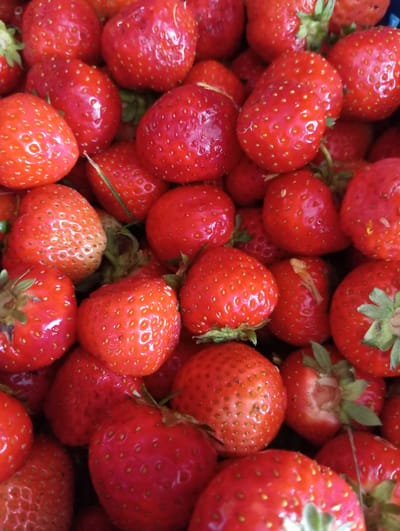 A close-up, overhead shot showcases a dense pile of ripe strawberries, filling the entire frame. The berries are a vibrant red, with visible seeds dotting their surfaces. Some strawberries have green stems or remnants of leaves attached. The lighting is even, highlighting the glossy texture of the fruit. The scene is devoid of any discernible environmental context, suggesting it may be an indoor setting or a tightly packed container. There are no people, actions, or interactions depicted. This image does not provide any information about a specific location in the city of Hlynytsi, Ukraine.