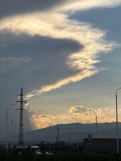 The image depicts a dramatic sky with heavy, dark clouds contrasted by bright, sunlit edges. A powerful electrical transmission tower stands silhouetted against this sky on the left. In the foreground, there is lush green vegetation and what appears to be a road with streetlights. Beyond the vegetation, vehicles, including a white van or minibus, are visible. In the background, a range of mountains forms the horizon, partially obscured by a hazy atmosphere. The lighting suggests either sunrise or sunset, casting a warm glow on the clouds and mountain peaks. The overall impression is one of a semi-urban or peri-urban environment on the outskirts of Dushanbe, Tajikistan, where infrastructure and natural landscapes converge under a striking sky.