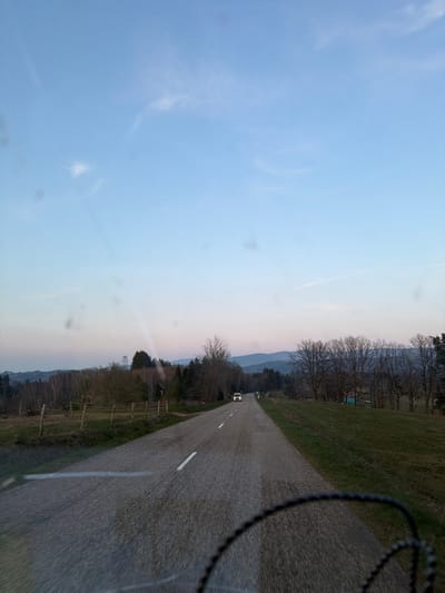 The image depicts a rural two-lane asphalt road in Le Saulcy, France, under a twilight sky, viewed from inside a vehicle. The road extends into the distance with a dashed white centerline. On the left, a grassy field bordered by a low wooden fence is visible, followed by a mix of bare deciduous and evergreen trees. A metal lattice tower stands amidst the trees on the left, and a small, dark structure is partially visible behind the fence. On the right, a grassy embankment slopes down to a treeline composed primarily of bare deciduous trees. Distant dark blue hills or mountains are visible on the horizon. A single white vehicle, possibly a van or SUV, with its headlights illuminated, is situated in the far distance on the road, facing away from the viewer. The sky exhibits a gradient from light blue at the zenith to a pinkish-orange hue near the horizon, with sparse cirrus clouds. Multiple circular smudges are present across the image, consistent with a camera lens or windshield. A portion of a dark, textured object, possibly a steering wheel or dashboard, is visible in the bottom right foreground. No individuals are discernible.