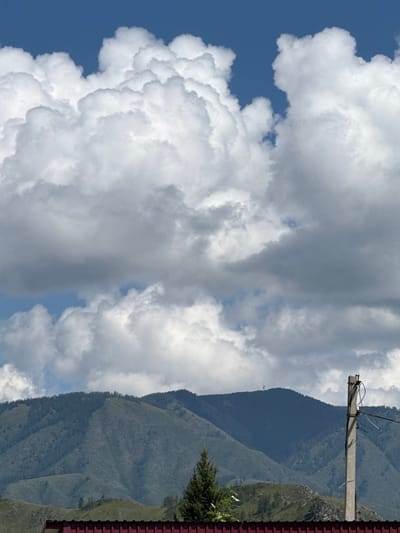 The image shows a rural landscape with a backdrop of mountains and a dramatic sky filled with large, fluffy white clouds against a deep blue sky. In the foreground, a red-tiled roof is visible. To the right of the frame, a concrete utility pole with wires extends upwards, partially obscuring the mountain range. The mountains are covered in sparse vegetation and appear to be hilly rather than sharply peaked. A solitary evergreen tree stands in front of the mountains, to the left of the utility pole. The scene suggests a remote, possibly mountainous region, consistent with the general geography of Onguday, Russia, which is located in the Altai Republic, known for its mountainous terrain. No people or specific activities are depicted in the image.