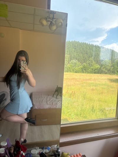 The media file is a vertical, reflected image taken indoors, looking out a window. The scene captured is a young woman in a light blue dress and black high heels, taking a selfie in a mirror. She is positioned on the right side of the frame, with her body angled and her right leg extended. Her dark hair falls around her shoulders as she holds up a smartphone to take the picture.
The mirror reflects the room she is in, showing a bed with a patterned cover in the background, and a dresser or cabinet. To the left of the woman's reflection, a light fixture with three woven lampshades hangs from the wall. A fan is visible at the bottom of the frame.
The window reveals an outdoor scene. A lush green field, transitioning to a denser green forest, stretches out under a bright blue sky with scattered white clouds. The time of day appears to be daytime, likely late morning or early afternoon, given the bright sunlight. The outdoor scenery suggests a rural or semi-rural location, consistent with the provided location context of Prodanovtsi, Bulgaria. The overall mood conveyed is casual and perhaps playful, as the woman poses for her selfie. There is no visible text in the image.