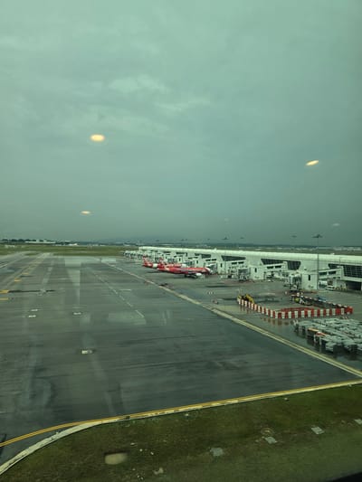 An elevated view captures an airport tarmac and terminal from what appears to be a window, possibly inside a passenger lounge or jet bridge. The sky is overcast and greyish-green, with two prominent, bright, oval reflections from interior lights visible in the window.

Numerous red AirAsia aircraft are parked along the modern airport terminal building. On the aircraft fuselages, the text "AirAsia.com" and "AirAsia" is clearly visible. On one particular plane, the word "CAMBODIA" is also discernible. The tarmac is a dark asphalt with various white and yellow ground markings; certain areas appear wet, suggesting recent rain. Ground support equipment, including items with red and white diagonal stripes and a yellow vehicle, is positioned near the aircraft and terminal.

To the right of the tarmac, a strip of vibrant green grass extends, featuring several square access covers embedded within it. The terminal building itself is characterized by its white, angular structures and large windows. The scene depicts typical daytime airport operations under cloudy weather conditions, consistent with a major regional hub like Kuala Lumpur International Airport (KLIA) in Sepang, Malaysia. No explicit emotions are conveyed in this functional, industrial setting.