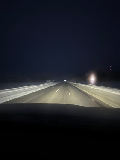 A nighttime view down a multi-lane highway illuminated by overhead lights. The dark sky on the left contrasts with the bright road stretching into the distance. A dark barrier or wall occupies the right side of the frame. The perspective is angled, giving a slightly distorted view of the road.