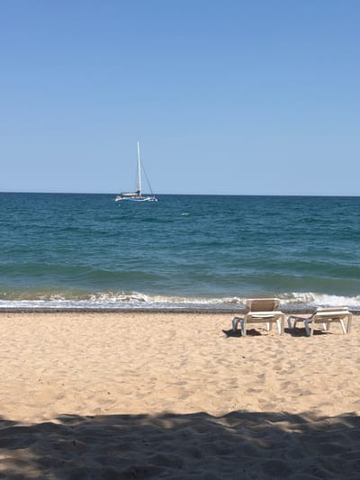 The image depicts a beach scene in Mont-roig del Camp, Spain. The scene includes the clear blue sky meeting the deep blue sea, which appears to have a sailboat. The waves crash gently on the sandy beach where there are four unoccupied beach chairs. The right side of the frame features a patch of beach shaded by what appears to be foliage.