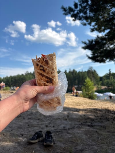 A person holds a partially eaten shawarma wrapped in plastic in their left hand. The shawarma has grill marks and visible filling. The background is a sandy area with sparse vegetation, and a body of water with beach umbrellas and people in the distance. A line of trees forms the horizon. The sky is blue with scattered white clouds. A pair of dark athletic shoes are on the ground in the foreground. The image appears to be taken on a sunny day.