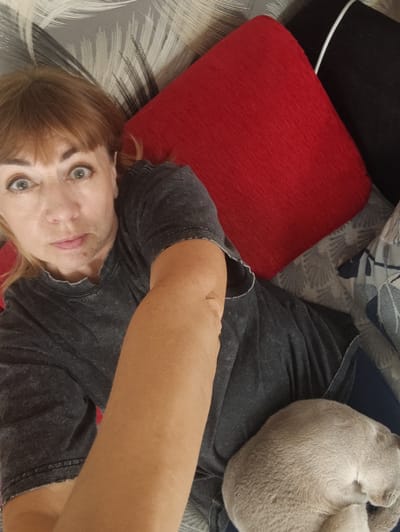 The image shows a woman and a cat lying on a sofa. The woman is in the foreground, facing the camera with her eyes wide open. She is wearing a dark grey t-shirt. Her right arm is extended towards the camera, obscuring part of the image. A light grey cat is curled up on the woman's lap, with its head tucked in. The sofa is covered with patterned cushions, including a large red one and a black one. The background shows a patterned cushion or blanket. The scene appears to be indoors. The location of Gvardeysk, Russia is not depicted or evident in the image.