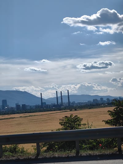The image depicts a panoramic view of Kazichene, Bulgaria, under a partly cloudy sky. In the foreground, a metal guardrail separates the viewer from a field of dry, golden crops. Behind the field, a city skyline is visible, with numerous buildings and industrial structures, including several tall smokestacks from a power plant. In the distance, a mountain range forms the backdrop, with a prominent television tower on one of the peaks. The overall scene suggests an industrial or semi-urban area, with elements of nature present in the fields and mountains. There are no people or animals visible in the image.