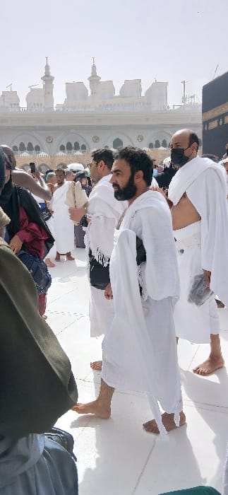 This image depicts a crowded outdoor scene within the Grand Mosque (Masjid al-Haram) complex in Makkah Al Mukarramah, Saudi Arabia. The iconic Kaaba, partially visible on the far right, is draped in black cloth adorned with gold calligraphy. In the background, the multi-story, light-colored architecture of the Grand Mosque rises under a bright sky, featuring numerous arched windows, balustrades, and multiple minarets.
The immediate foreground and midground are populated by a diverse group of pilgrims. Several men are visible, predominantly barefoot and dressed in the white two-piece Ihram garments worn for Hajj or Umrah. A central male figure, with a beard, walks from left to right, his gaze directed forward. He wears Ihram and has a dark object tucked under his left arm. To his immediate right, another male pilgrim in Ihram, also barefoot, wears a black face mask and holds a dark, rectangular object in his left hand. To the left of the central figure, a third male pilgrim in Ihram carries a light-colored, fan-shaped object in his right hand. Numerous other individuals, including both men in Ihram and women in darker, traditional attire, are observable in various states of movement across the white, reflective tiled floor of the courtyard. The lower-left corner shows partial views of dark green and blue fabric, indicative of the photographer's clothing or an adjacent pilgrim. The scene conveys a large congregation performing religious observances in the holy city.
