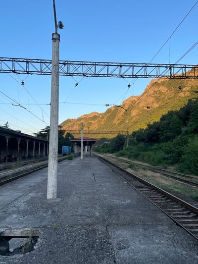 This is a vertical, slightly low-angle shot of a train platform and tracks next to a steep, forested mountain. The scene is set in Mtskheta, Georgia. The time of day appears to be late afternoon or early evening, as the sun casts a warm, golden light on the mountain face, illuminating the rugged terrain and the dense green foliage. The sky is a clear, bright blue, suggesting fair weather.
In the foreground, the concrete platform is worn and weathered, showing signs of age and damage, with a large hole revealing some debris underneath. A tall, white pole with a lamp fixture stands prominently on the platform, casting a shadow. To the left of the platform, a lattice-like metal structure supports overhead electrical wires, typical of a railway line. The tracks themselves are visible, running parallel to the platform and disappearing into the distance.
In the middle ground, there is a small, covered structure that resembles a bus stop or a small station building, with a red roof. A blue portable toilet is parked nearby. The mountain rises steeply behind the tracks and the building, its slopes covered in trees that transition from dark green to a sun-drenched golden hue on the upper parts.
The overall mood of the image is one of quiet desolation and natural beauty. The abandoned-looking platform and the vastness of the mountain suggest a sense of remoteness. There is no visible text in the image.
