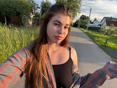 A young woman with long, brown hair is captured in a selfie, outdoors on a paved road. She is wearing a black tank top and an open plaid shirt in shades of grey, red, and blue. Her hair is styled naturally and falls around her shoulders. She is looking directly at the camera with a neutral expression, her lips slightly parted. The background suggests a rural setting with houses and trees visible on either side of the road. The sun is low in the sky, casting long shadows and illuminating the scene with warm, golden light, indicating it is likely late afternoon or early evening. The sky is partly cloudy. The image is oriented sideways.