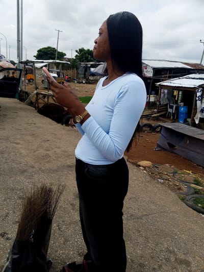 A young Black woman stands on a paved road, facing right, looking at her phone. She wears a light blue long-sleeved shirt and black pants, with a gold watch on her left wrist. In her left hand, she holds a pink smartphone. A large bunch of dried grass in a black plastic bag stands on the ground next to her left leg. Behind her, various stalls and structures with corrugated metal roofs are visible, along with parked vehicles, including a yellow tricycle. The ground is a mix of dirt and gravel, with discarded tires and other debris scattered around. The sky is overcast. The image appears to be taken in Pasere, Nigeria.