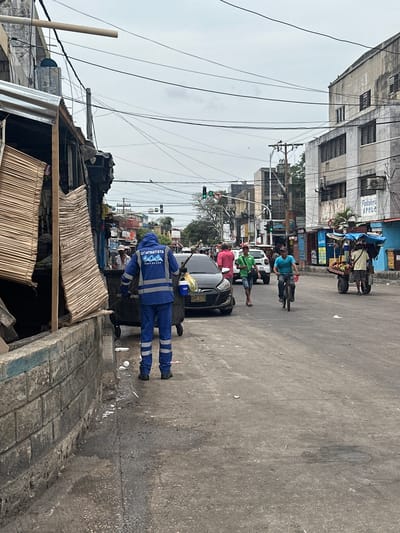 This image captures a street scene in Perímetro Urbano Barranquilla, Colombia. In the foreground, a sanitation worker in a blue uniform with reflective stripes is standing next to a large trash receptacle, appearing to be sweeping or attending to waste. To the left, a weathered building with a partially visible wooden structure and bamboo blinds lines the street. The street itself is asphalted and shows signs of wear and tear.
Further down the street, traffic is visible, including a black sedan with its license plate partially obscured, a cyclist in a teal shirt, and a cart laden with produce being pulled by a person in a light-colored shirt. Several other pedestrians and buildings, some with awnings and signage indicating businesses, are present in the background. Traffic lights are visible overhead, suggesting an intersection. The sky is overcast, contributing to the overall muted lighting of the scene. The environment appears to be a busy urban area, with a mix of commercial and residential structures.