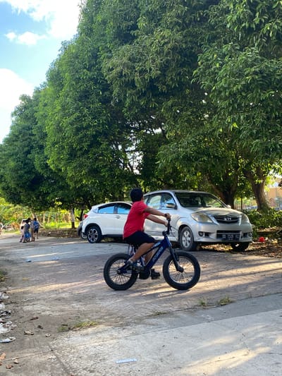The image depicts a boy riding a bicycle on a paved path in Ta', Indonesia. The boy, wearing a red shirt and black shorts, is in the foreground, with the rear wheel of his bike prominently visible. In the background, two cars are parked: a silver SUV and a white SUV. Several trees with dense green foliage line the right side of the image, casting shadows on the ground. A group of people, possibly children, are visible in the distance to the left, near some vegetation. The scene appears to be outdoors, during daylight hours, with a bright sky and scattered clouds. The ground is a mix of paved surfaces and dirt, with some debris present.