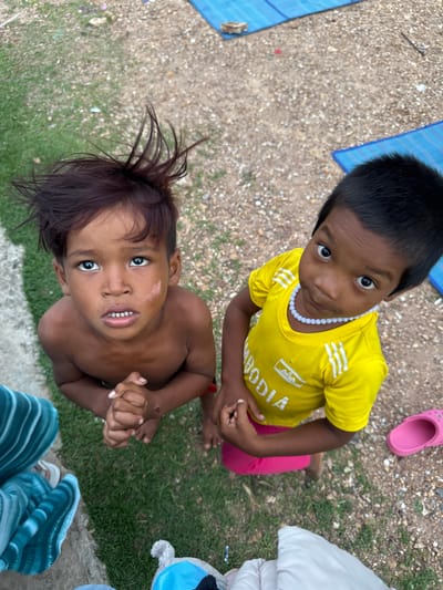 Two young children, a boy and a younger boy, are captured from an overhead perspective in Siem Reap, Cambodia. The older boy, on the left, has dark, windswept hair and is shirtless, with his hands clasped in front of him. He has a light-colored mark on his right cheek. The younger boy, on the right, wears a bright yellow t-shirt with white stripes on the sleeves and a white beaded necklace. His t-shirt has "CAMBODIA" printed on it in white lettering, along with a graphic of a building. He is also wearing bright pink shorts. Both children appear to be looking upwards, potentially at the photographer. The ground beneath them is a mix of dirt, pebbles, and sparse grass. In the background, two blue mats are visible on the ground, and a pink sandal lies to the right of the younger boy. To the left, a person's legs are visible, covered in a striped fabric. The scene suggests an outdoor setting, possibly a residential area or a public space in Siem Reap.