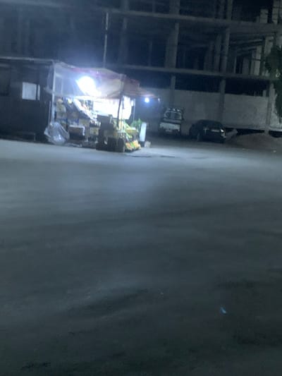 A nighttime scene shows a street with a small market stall illuminated by bright lights. The stall displays produce and packaged goods. Behind the stall, a building under construction with exposed scaffolding is visible. A vehicle is parked on the street further back. The scene appears to be shot from a slightly elevated position, and the image is rotated counter-clockwise. The street itself dominates much of the right side of the frame.