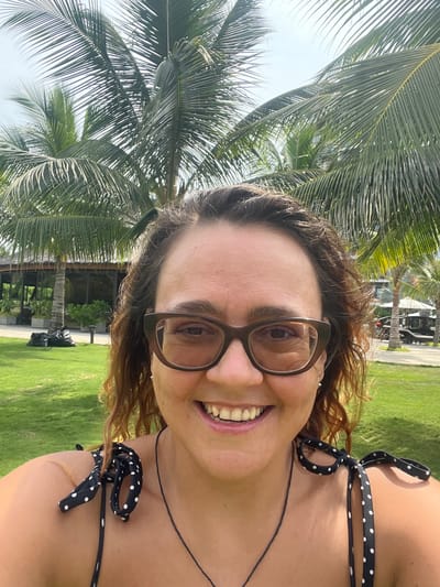 A woman is taking a selfie in Cam Lam, Vietnam, with palm trees in the background. She is wearing glasses and a black and white polka dot bikini top. She is smiling and appears to be outdoors on a sunny day. The background shows a grassy area, some buildings, and more palm trees.
