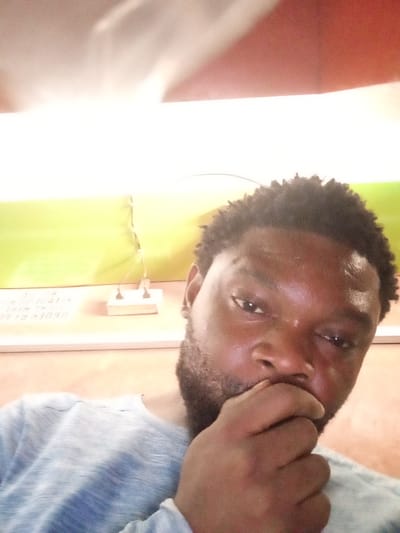 An indoor, eye-level selfie shows an adult male of apparent Nigerian ethnicity, positioned slightly off-center to the right. He possesses short, dark, textured hair and a dark beard. His right hand is partially covering his mouth and lower chin. He is wearing a light-colored, possibly grey or light blue, long-sleeved garment. The background consists of a bright, overexposed light source at the top, transitioning to a light green horizontal surface, and then a lower light brown/orange surface. On the green surface, to the left of the male's head, a small white object with two dark, vertical components is visible. Obscured, mirrored text, including the fragment "ORDER," is faintly visible on the green surface behind the subject. The setting is identified as an interior location within the city of Magboro, Nigeria.