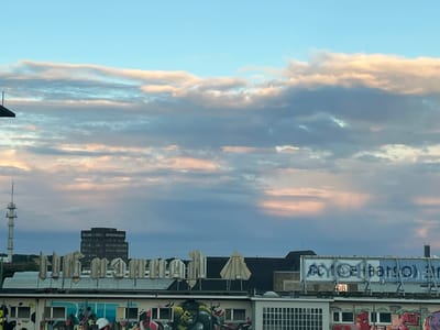This is a rooftop view of the city of Mönchengladbach, Germany, with a focus on graffiti and a distant office building. In the foreground, a row of large, white, three-dimensional letters is mounted on a building. Some of these letters are damaged or incomplete. Below this, the building's facade is covered in colorful graffiti, including a prominent depiction of the Incredible Hulk. To the left of the building with the letters, a tall, slender communications tower is visible. Further in the background, a dark, rectangular office building stands against a cloudy sky. The sky itself is a blend of blue, gray, and streaks of pink and orange, indicating a sunset or sunrise. The overall scene is urban and industrial, with a strong emphasis on street art.